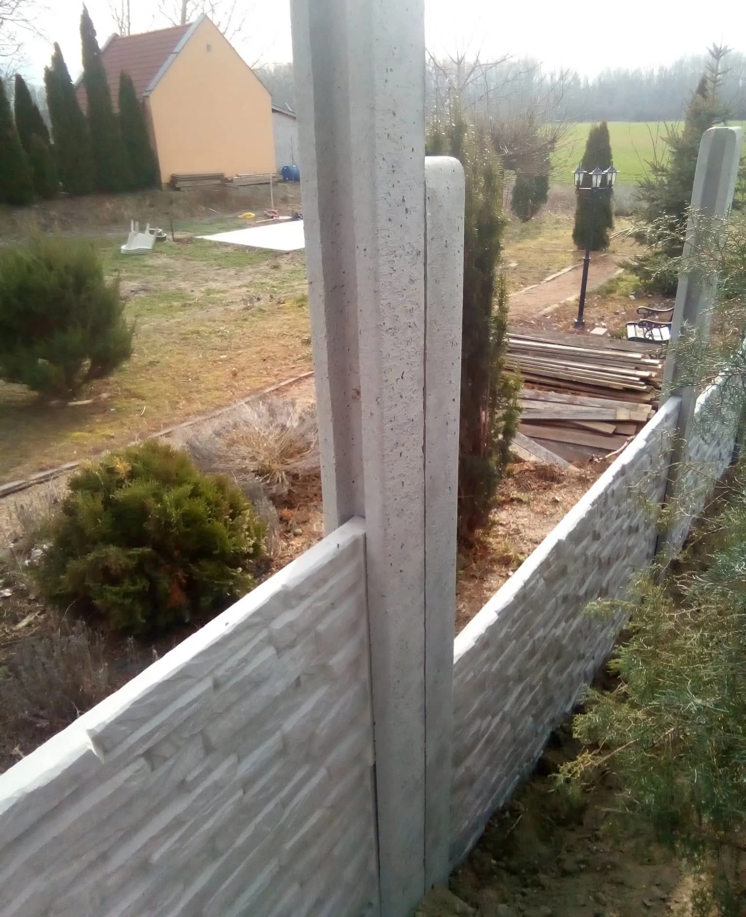 Kőmintás betonkerítés lejtős kertben: a mezők az oszlopoknál lépcsőzetesen követik a terepet